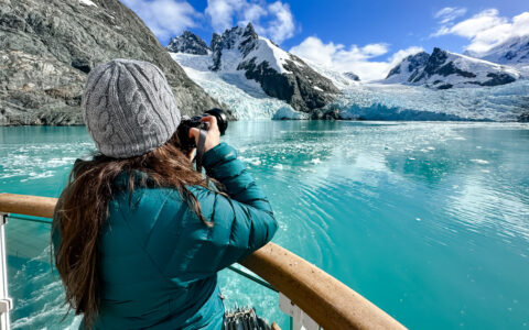 Woman in teal down coat & knitted hat holds camera over deck railing to photograph a blue glacier & snowy peaks in Antarctica.