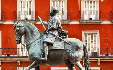 statue of soldier riding a horse with a weapon in hand infront of bright red building with white balconys