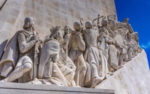 Stone statue on the side of a historic building showing men and women walking up stairs to the bright blue sky