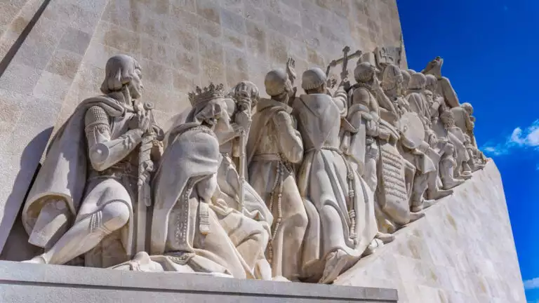 Stone statue on the side of a historic building showing men and women walking up stairs to the bright blue sky