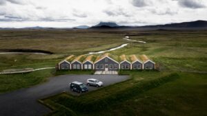 Aerial view of 6 slender grey buildings, 3 on each side of a wider one, with turf roofs & parking lot with 2 cars out front.