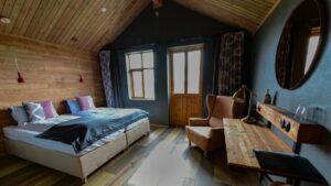 Modern guest room with double bed, wooden desk, wooden accent wall & ceiling, window, door, 3 teal walls, 2 peach armchairs & round mirror.