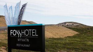 Outdoor hotel sign in dark brown with flags rising behind, advertises the Fosshotel Myvatn, with building in background & green hills in front.