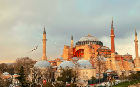 Turkish mosque with various gray domes, tall, pointed minarets & beige stone walls seen at dusk.