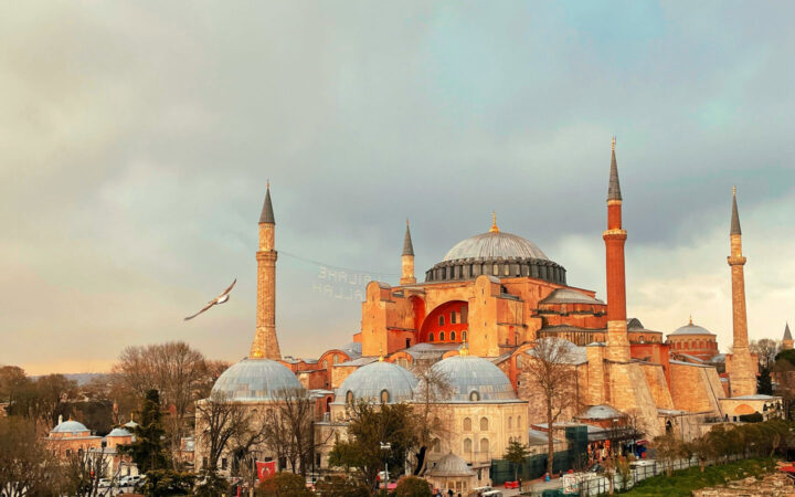Turkish mosque with various gray domes, tall, pointed minarets & beige stone walls seen at dusk.