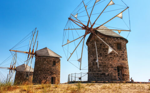 Historic windmills with round brick buildings as stands sit under bright blue sky in Greece.