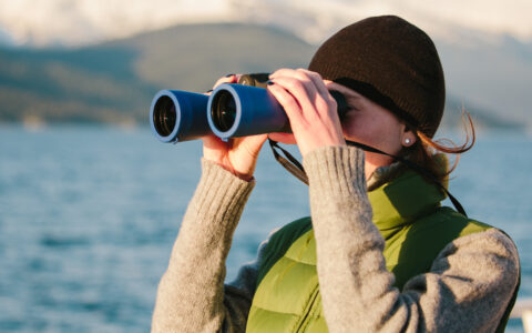 Woman in black knit cap & green puffy vest holds blue binoculars up to look at wildlife on a Gulf of Alaska cruise.