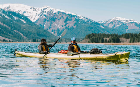 Tandem kayakers paddle a yellow boat in calm water with snowcapped peaks behind on a sunny day of a Gulf of Alaska cruise.