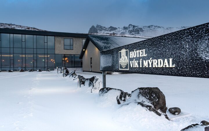 Exterior ground-level view of Iceland Hotel Vik i Myrdal in winter at dusk, with glass walls & illuminated black rectangular sign.