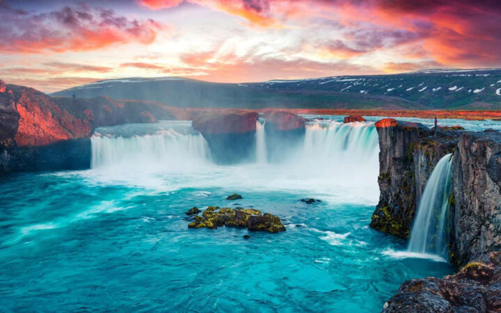 Huge Iceland waterfall sends water down rocky cliffs into a teal lagoon while a traveler stands at the edge at sunset.
