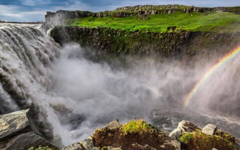 Huge waterfall sends rushing water down past lush green hillsides, with spray from the water creating a rainbow on the Iceland Adventure.