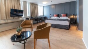Double bed in modern hotel room with view windows, flat screen TV & seating area with 2 padded tan chairs & small black coffee table.