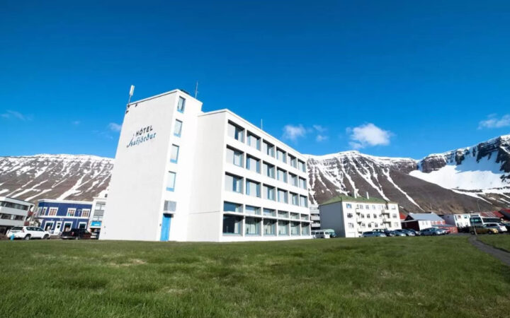 Exterior view of Iceland Hotel Isafjordur, a 4-story white building in the center of town, with green grass & mountains behind.