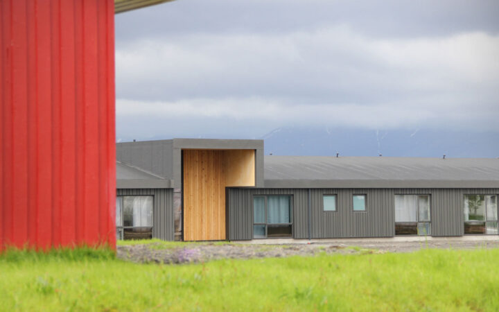 Red metal building & bright green grass front a modern gray building with wooden accent wall on a cloudy day in Iceland.