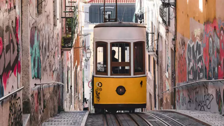 Yellow & white cable car climbs stone street between two graffitied buildings in Lisbon, Portugal, seen on the Med Feast cruise.