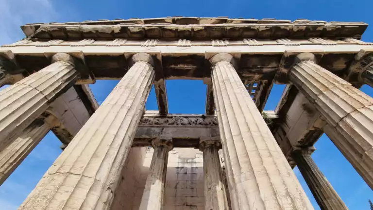 Looking up tall stone pillars holding an open, carved stone roof under a blue sky at the Acropolis in Athens, Greece.