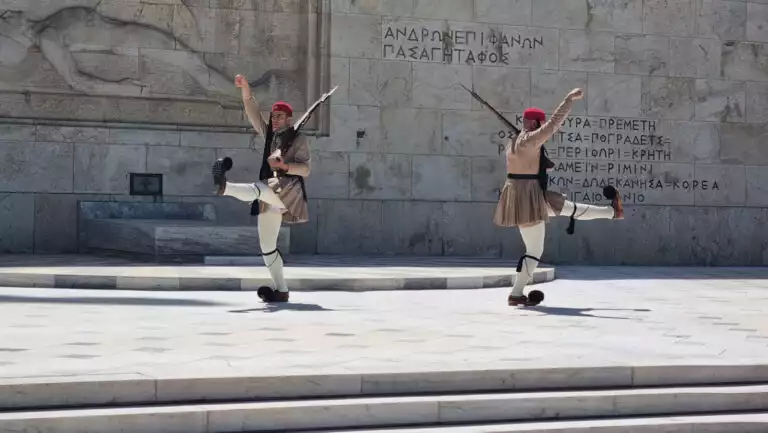 2 armed Greek guards in traditional garb wave arms & legs in a ceremonial changing of the guard atop a beige stone plaza.