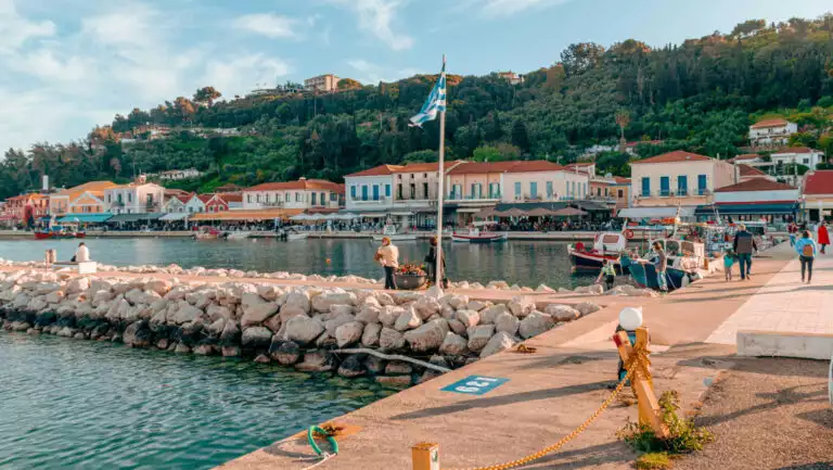 Beige stone seawall beside public dock & seaside village backed by green hills, seen on an Athens to Naples cruise.