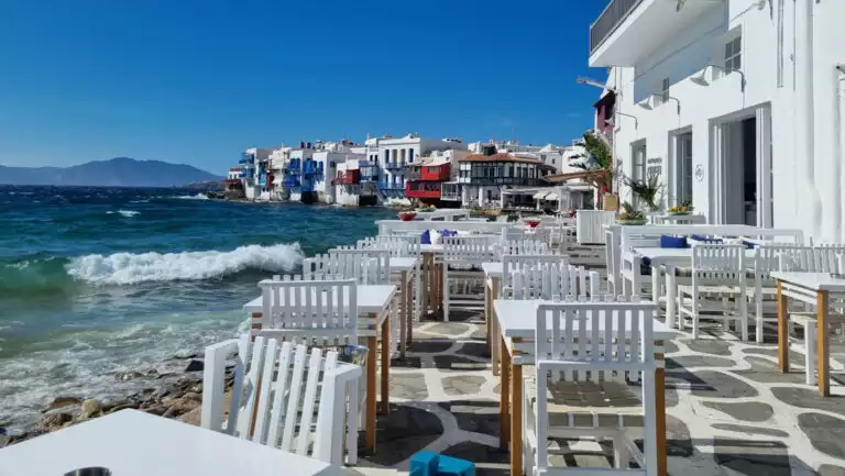 Empty whitewashed tables, chairs & restaurants sit seaside on a sunny day with turquoise waves crashing in Mykonos, Greece.