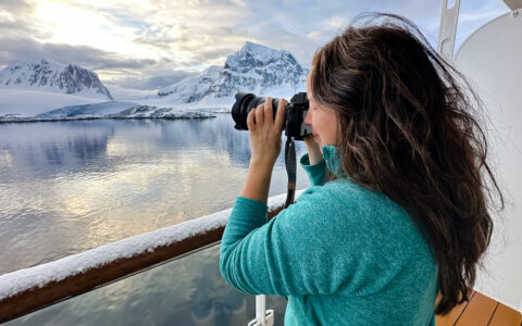 Woman in teal sweater stands on ship deck & photographs a white polar landscape in Antarctica during the Ross Sea Odyssey.