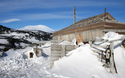 Rustic, historic wooden Shackleton hut sits among snow & ice by dark mountain under blue skies in Antarctica.