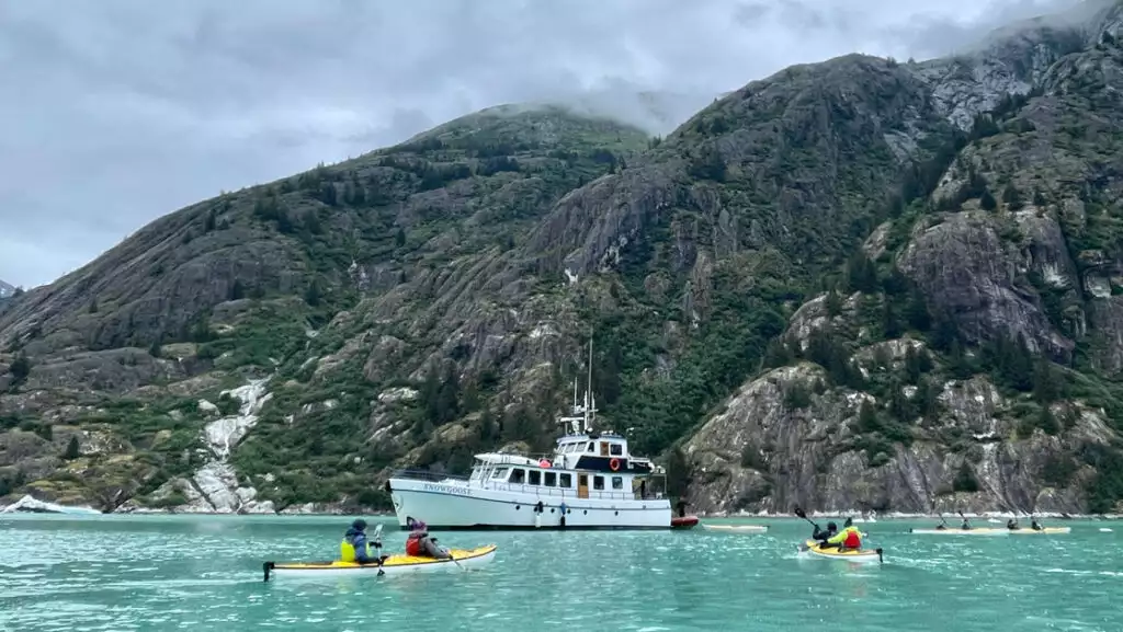 Snow Goose Alaska yacht sits in milky teal sea as guests paddle tandem yellow sea kayaks beside a rocky mountainside.