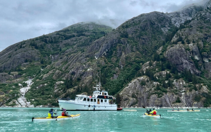 Snow Goose Alaska yacht sits in milky teal sea as guests paddle tandem yellow sea kayaks beside a rocky mountainside.