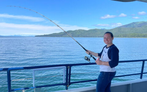 Woman stands behind blue railing on deck of the Snow Goose yacht in Alaska, holding a fishing rod & smiling on a sunny day.