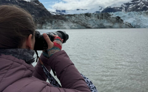 Woman in purple down jacket & fingerless gloves points a camera over water to an icy blue & black glacier in Alaska.