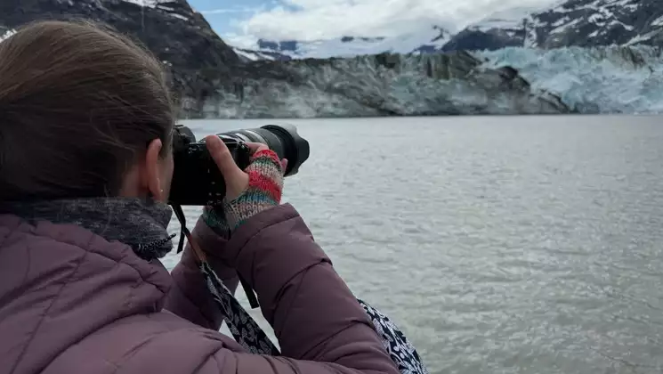 Woman in purple down jacket & fingerless gloves points a camera over water to an icy blue & black glacier in Alaska.