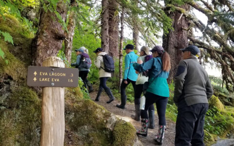 Small group of hikers walks a forested trail past a forest service sign for Lake Eva on a cloudy day in Alaska.