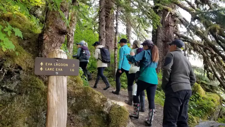 Small group of hikers walks a forested trail past a forest service sign for Lake Eva on a cloudy day in Alaska.