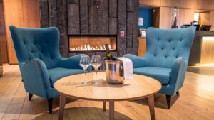 Reception lobby at Hotel Vik i Myrdal, with gas fireplace, wood flooring, 2 teal wingback chairs & wooden coffee table set for champagne service.
