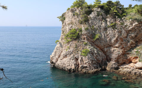 Turquoise sea spills past rocky outcrop & into small inlet with swimmers on a sunny day in Croatia.