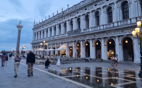 Travelers walk gray stone tile walkway beside long, Roman-inspired building at dusk in Venice, Italy.