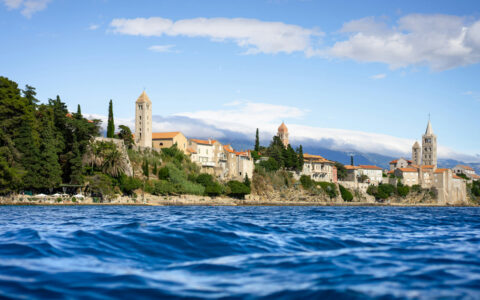 Stunning coastline of Croatia with historic beige stone buildings, terra cotta roofs, belltowers & green trees by blue sea.