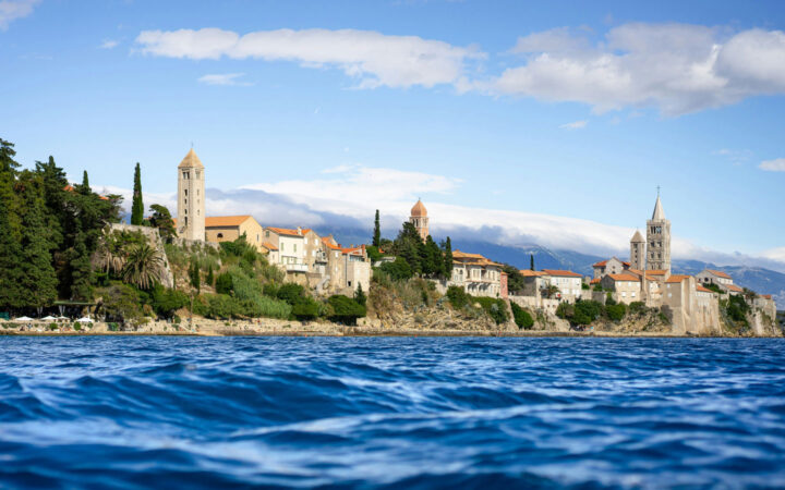 Stunning coastline of Croatia with historic beige stone buildings, terra cotta roofs, belltowers & green trees by blue sea.