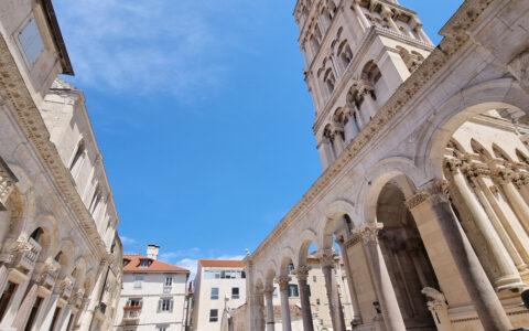 Looking up to tops of historic beige stone buildings with terra cotta roofs & a belltower, under blue sky in Croatia.
