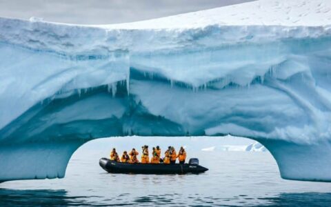 A zodiac boat is visible beneath an iceberg that's arching over the ocean, in antarctica