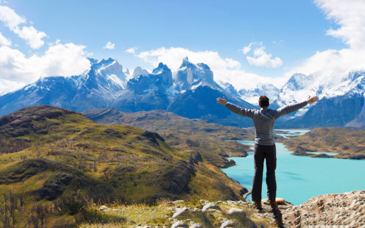 Man stands on grassy hilltop looking down on a turquoise lake with jagged mountain peaks in the background during the Essential Patagonia: Chilean Fjords & Torres del Paine Cruise.