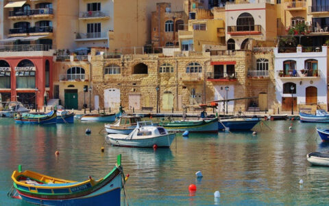Small, colorful wooden fishing boats sit in calm harbor by Mediterranean stone buildings on a sunny day.