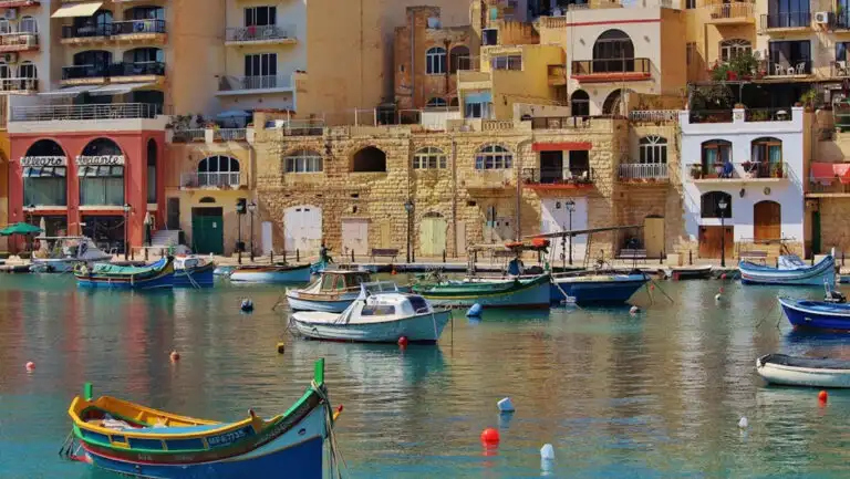 Small, colorful wooden fishing boats sit in calm harbor by Mediterranean stone buildings on a sunny day.