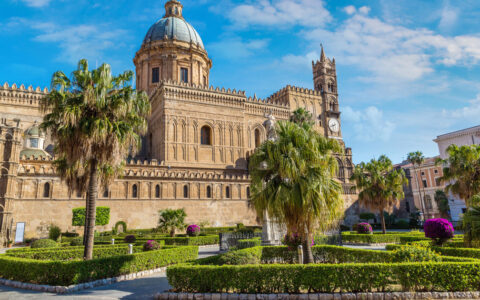 Mediterranean civic center with palm trees & manicured green hedges by tan brick building with blue dome under sunny skies.