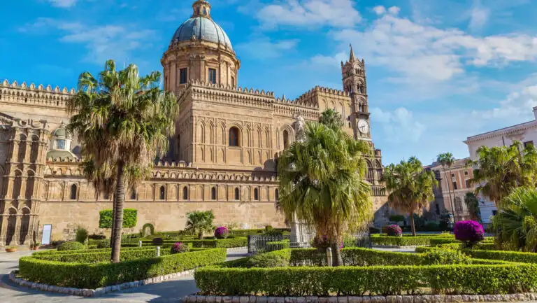 Mediterranean civic center with palm trees & manicured green hedges by tan brick building with blue dome under sunny skies.