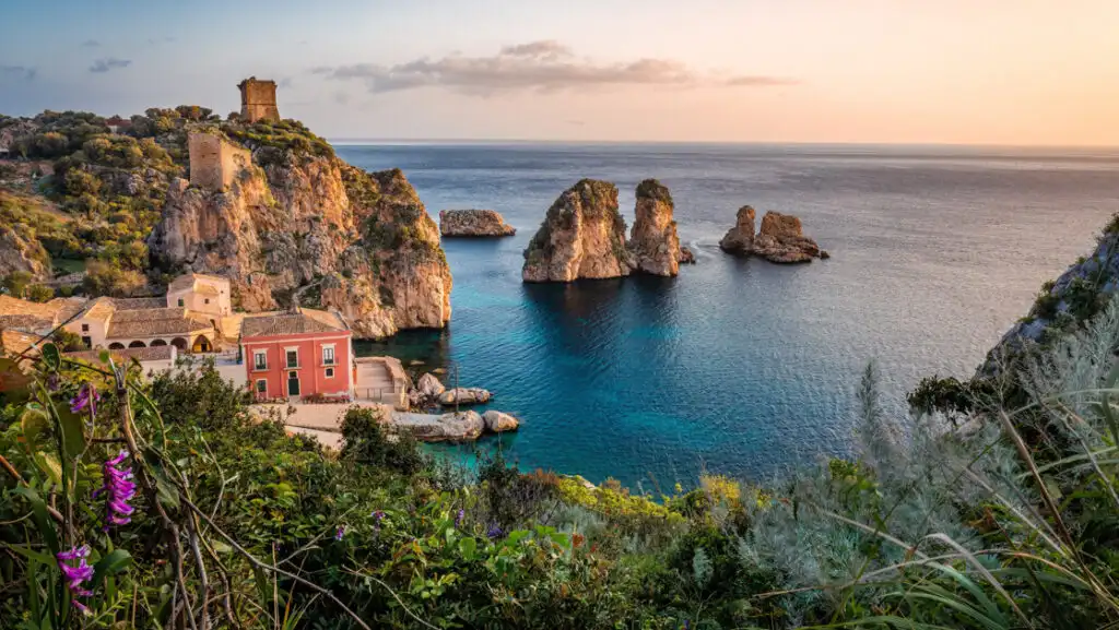 Sunset from rocky Sicilian coast with tan brick buildings beside teal sea & flowering cliffs.
