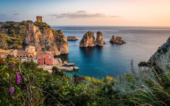 Sunset from rocky Sicilian coast with tan brick buildings beside teal sea & flowering cliffs.
