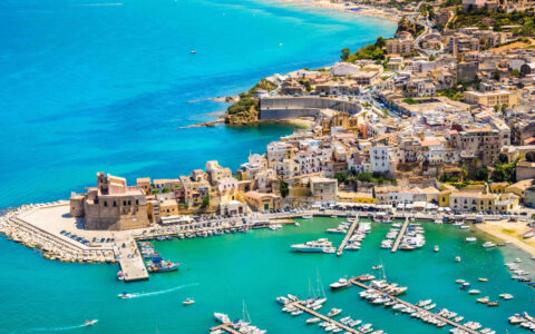 Aerial view of Mediterranean seaside city with boats in small harbor of teal sea, white-sand beach & seaside walk.