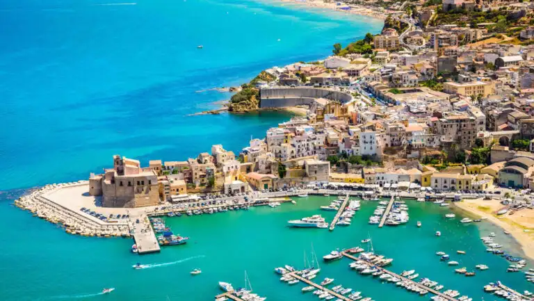 Aerial view of Mediterranean seaside city with boats in small harbor of teal sea, white-sand beach & seaside walk.