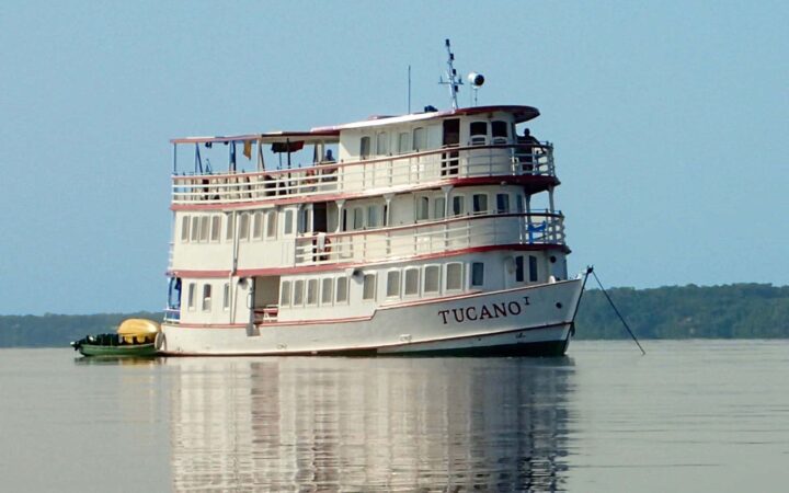 amazon small cruise ship sitting on calm water with a kayak tied to the back and jungle in the distance