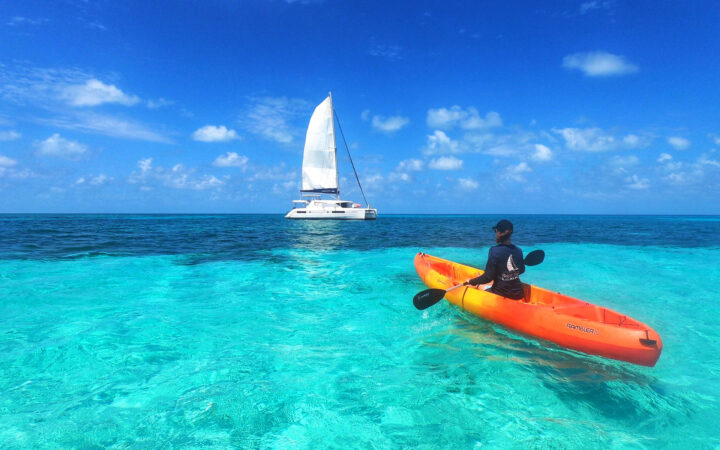 Woman paddles bright orange & yellow kayak in turquoise sea near a white catamaran under sunny skies on a Belize adventure.